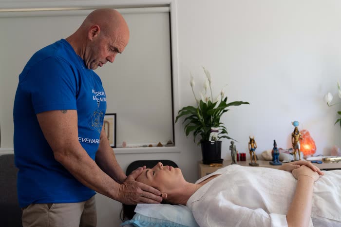 Steve standing at the head of the treatment table, holding a client's head as she lies down, fully clothed, at the start of a reiki session.